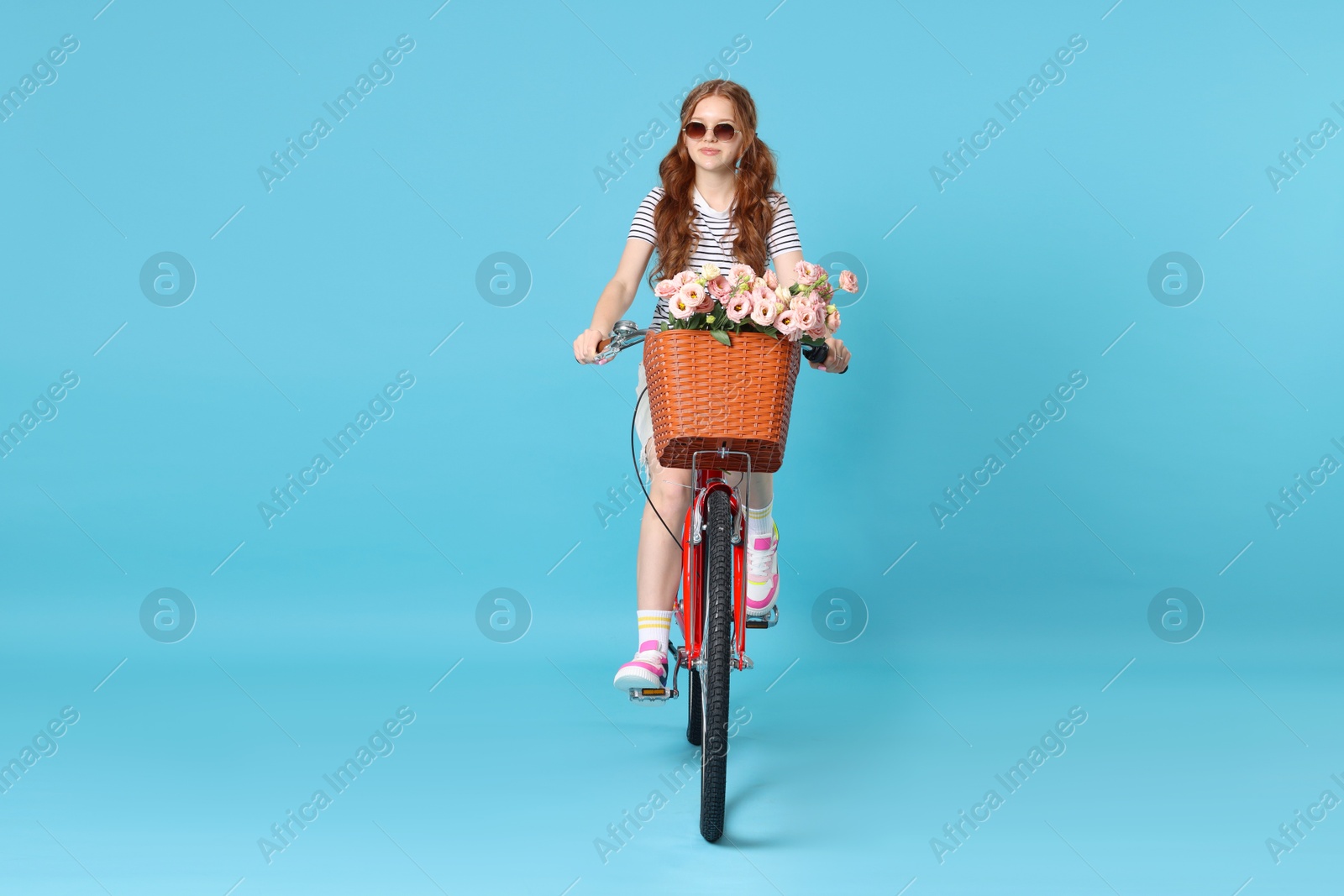 Beautiful teenage girl in sunglasses riding bicycle with basket of flowers against light blue background Photo of Beautiful teenage girl in sunglasses riding bicycle with basket of flowers against light blue background