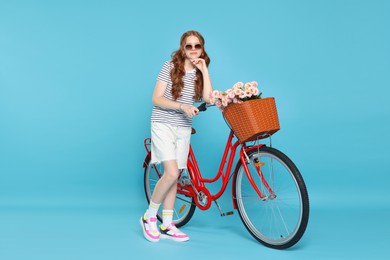 Photo of Beautiful teenage girl in sunglasses and bicycle with basket of flowers against light blue background. Space for text