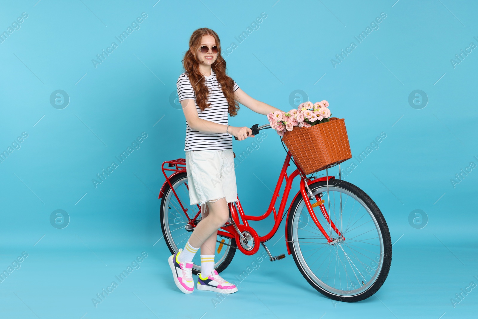 Smiling teenage girl in sunglasses and bicycle with basket of flowers against light blue background Photo of Smiling teenage girl in sunglasses and bicycle with basket of flowers against light blue background