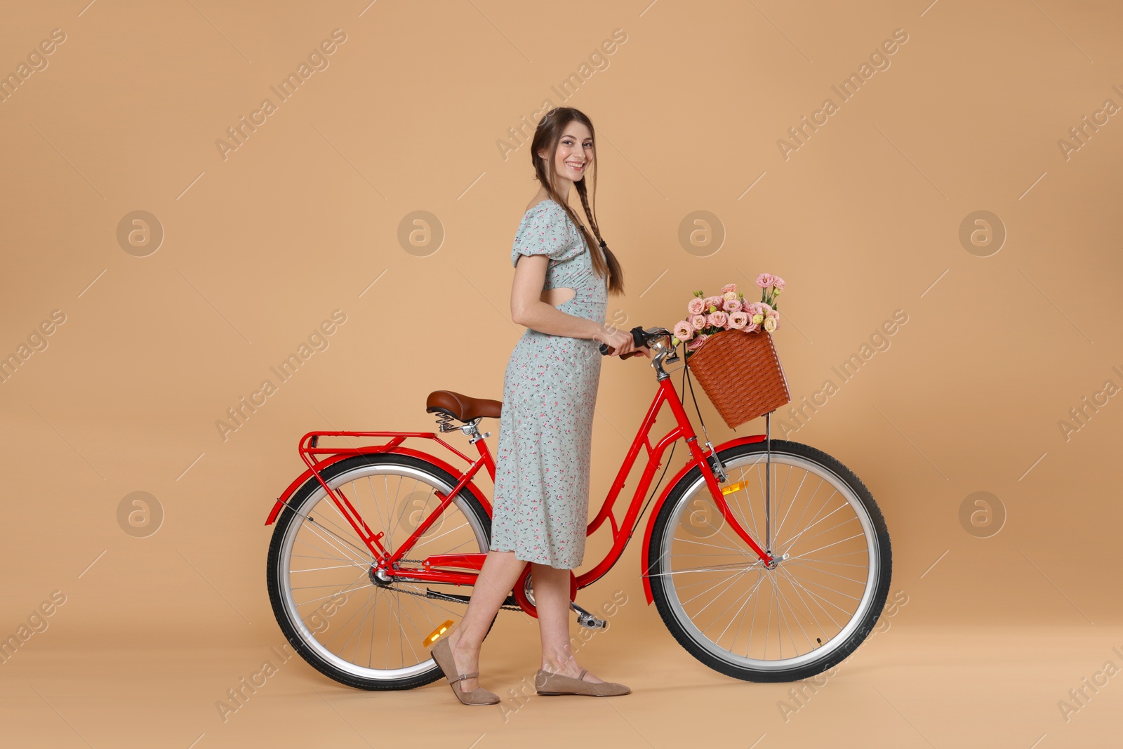 Smiling woman in dress and bicycle with basket of flowers on beige background Photo of Smiling woman in dress and bicycle with basket of flowers on beige background