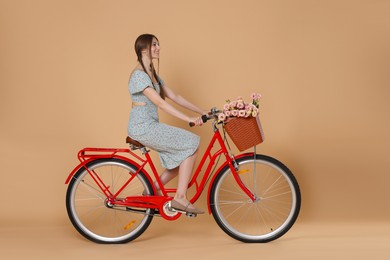 Photo of Smiling woman in dress riding bicycle with basket of flowers on beige background