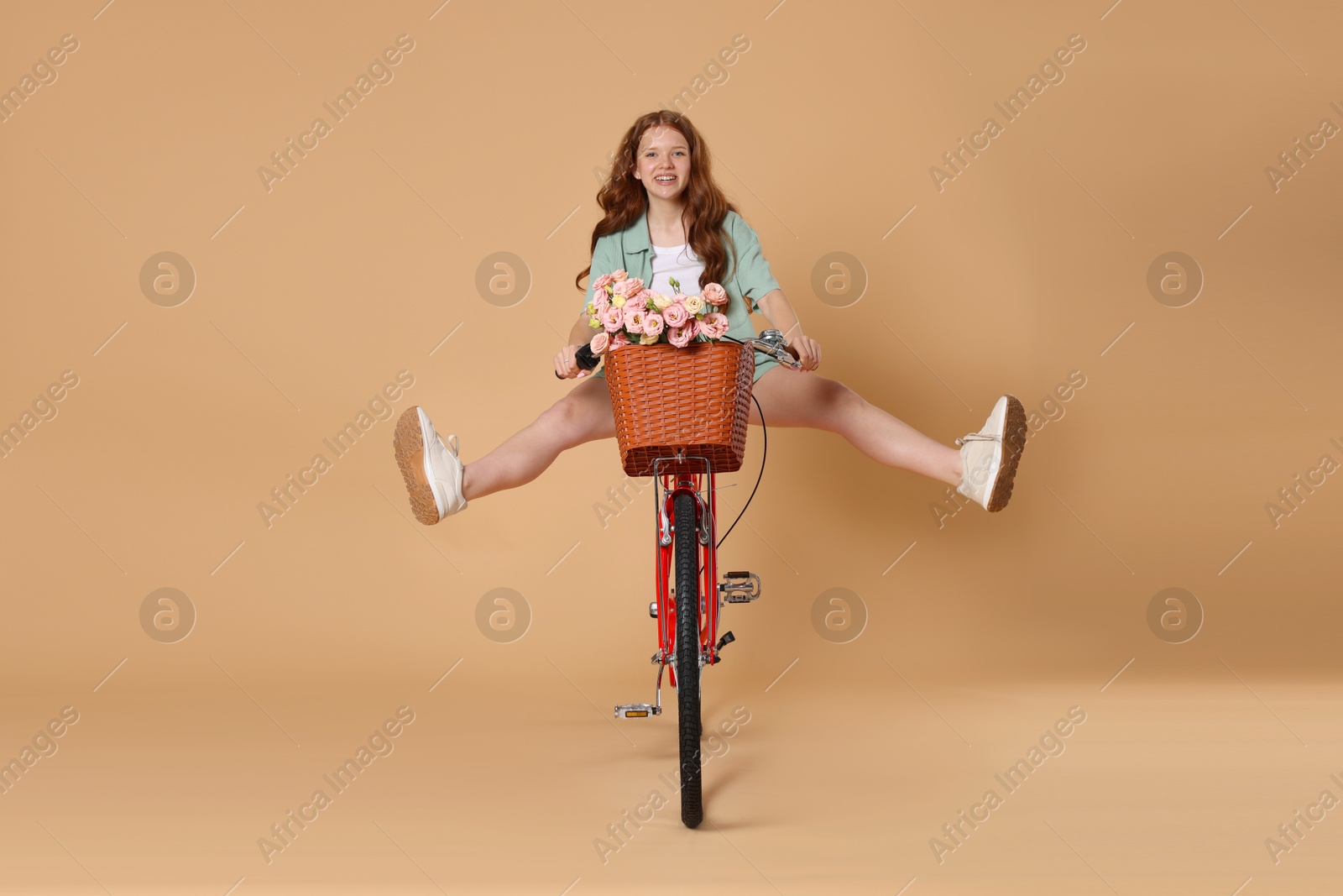 Smiling teenage girl riding bicycle with basket of flowers against beige background Photo of Smiling teenage girl riding bicycle with basket of flowers against beige background