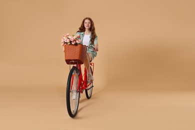 Photo of Beautiful teenage girl riding bicycle with basket of flowers against beige background. Space for text