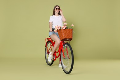 Photo of Smiling woman in sunglasses riding bicycle with basket of flowers on green background