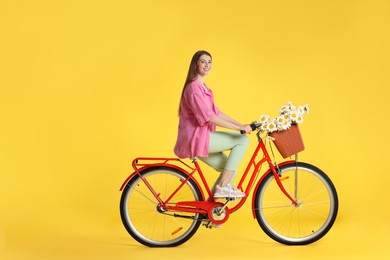 Photo of Smiling woman riding bicycle with basket of flowers against yellow background. Space for text