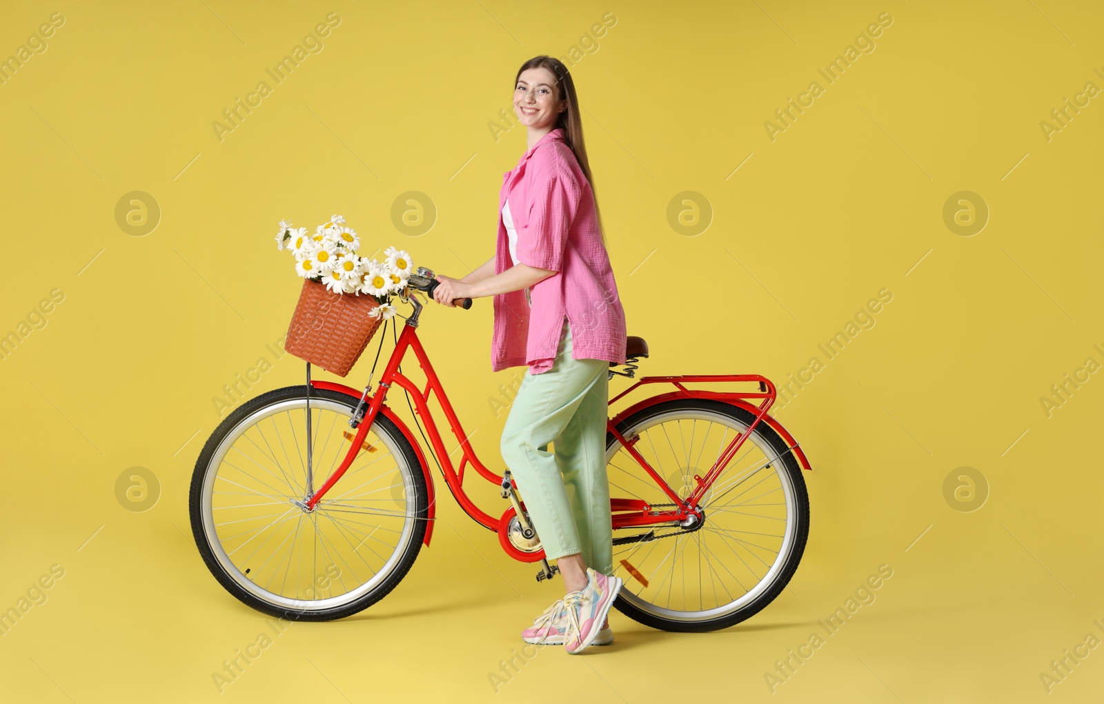 Smiling woman and bicycle with basket of flowers against light green background Photo of Smiling woman and bicycle with basket of flowers against light green background