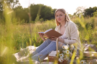 Photo of Woman with bouquet of wildflowers reading book on blanket in meadow