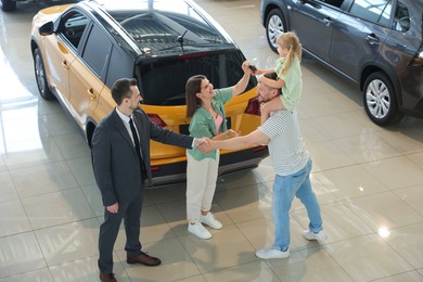 Photo of Salesman shaking hands with happy family near new car in salon