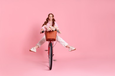 Photo of Beautiful teenage girl riding bicycle with basket of flowers on pink background