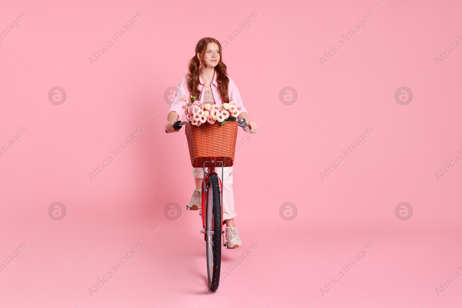 Photo of Beautiful teenage girl riding bicycle with basket of flowers on pink background