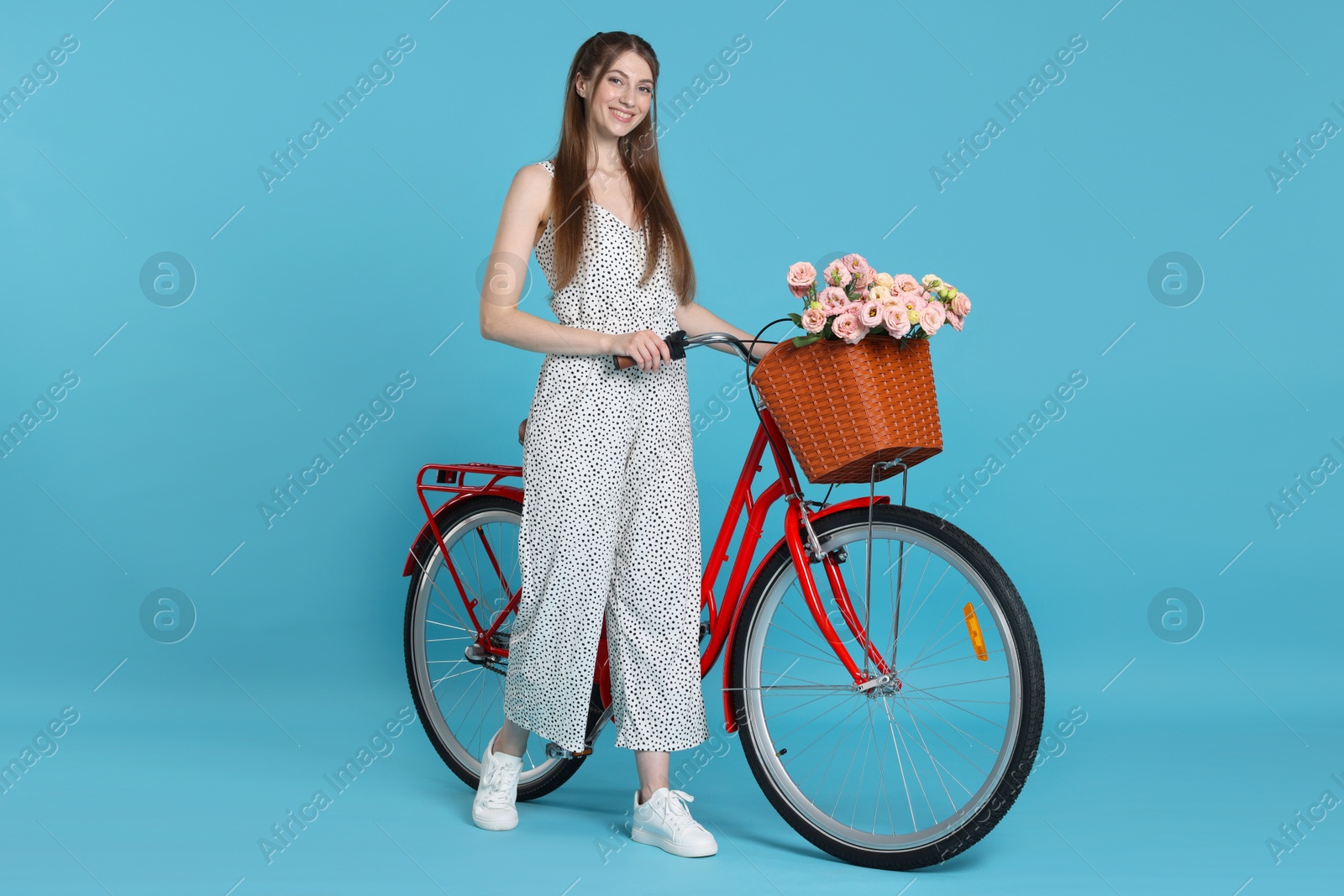Smiling woman and bicycle with basket of flowers against light blue background Photo of Smiling woman and bicycle with basket of flowers against light blue background