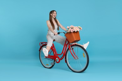 Photo of Happy woman riding bicycle with basket of flowers against light blue background