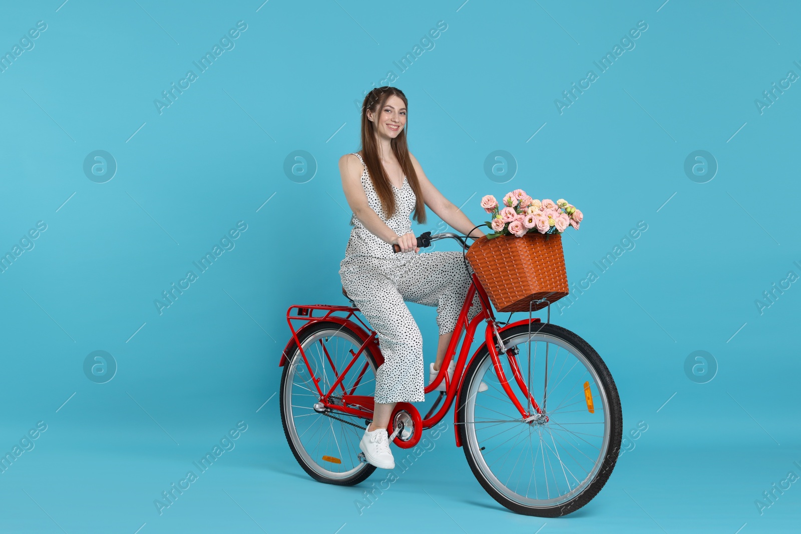 Smiling woman riding bicycle with basket of flowers against light blue background Photo of Smiling woman riding bicycle with basket of flowers against light blue background