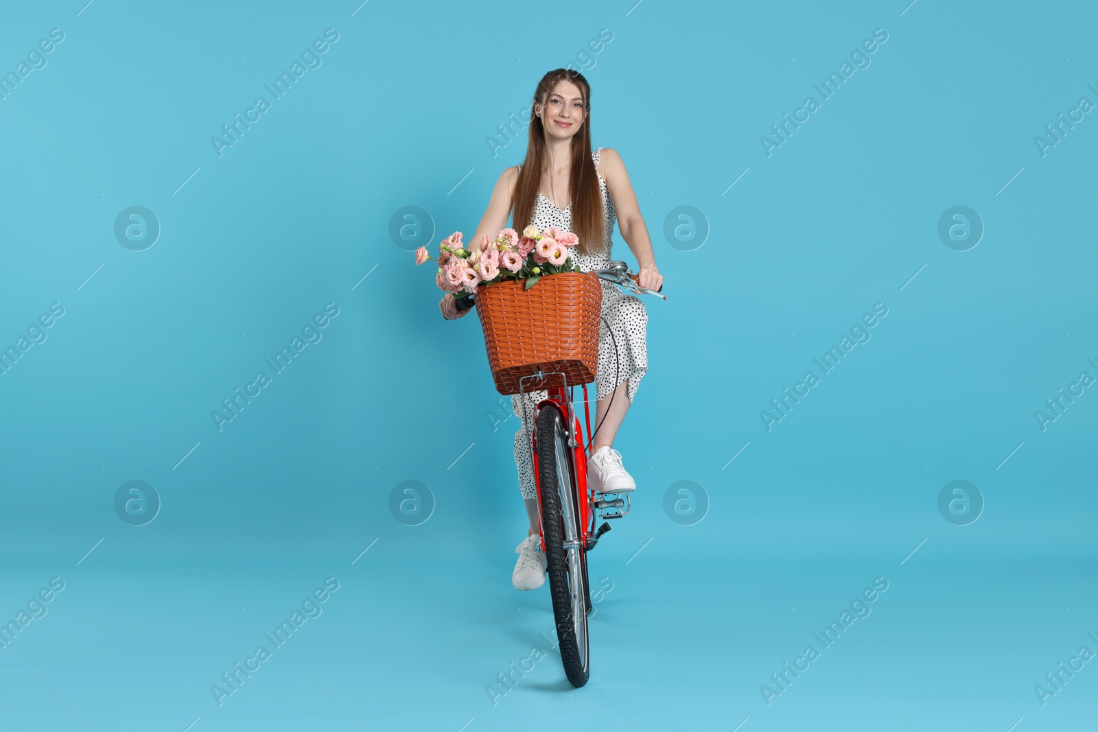 Beautiful woman riding bicycle with basket of flowers against light blue background Photo of Beautiful woman riding bicycle with basket of flowers against light blue background