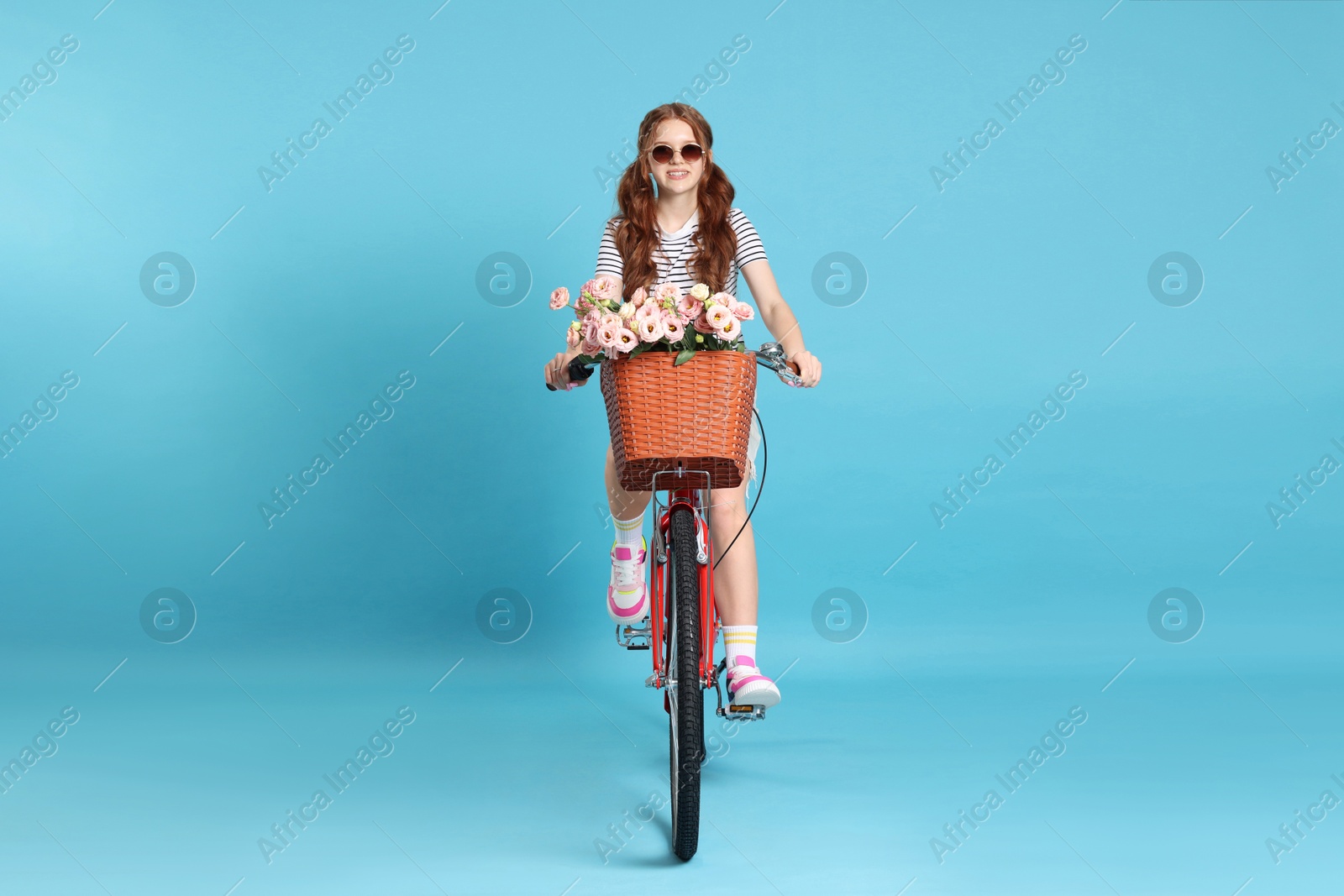 Smiling teenage girl in sunglasses riding bicycle with basket of flowers against light blue background Photo of Smiling teenage girl in sunglasses riding bicycle with basket of flowers against light blue background