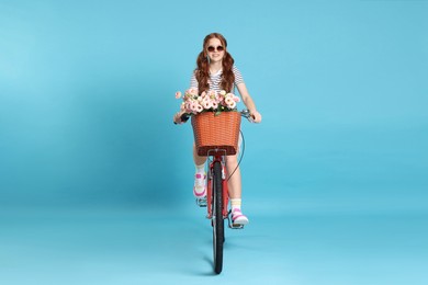 Photo of Smiling teenage girl in sunglasses riding bicycle with basket of flowers against light blue background