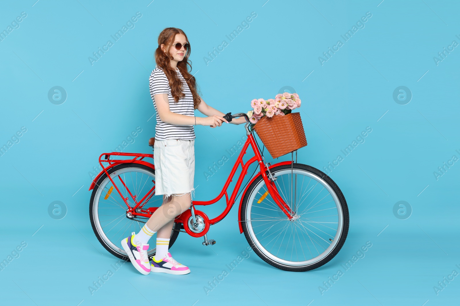 Beautiful teenage girl in sunglasses and bicycle with basket of flowers against light blue background Photo of Beautiful teenage girl in sunglasses and bicycle with basket of flowers against light blue background