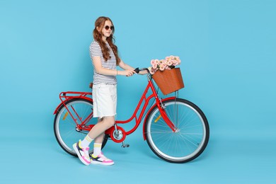 Photo of Beautiful teenage girl in sunglasses and bicycle with basket of flowers against light blue background