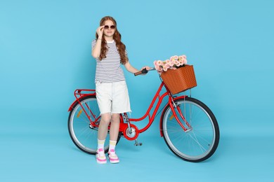 Photo of Beautiful teenage girl in sunglasses and bicycle with basket of flowers against light blue background