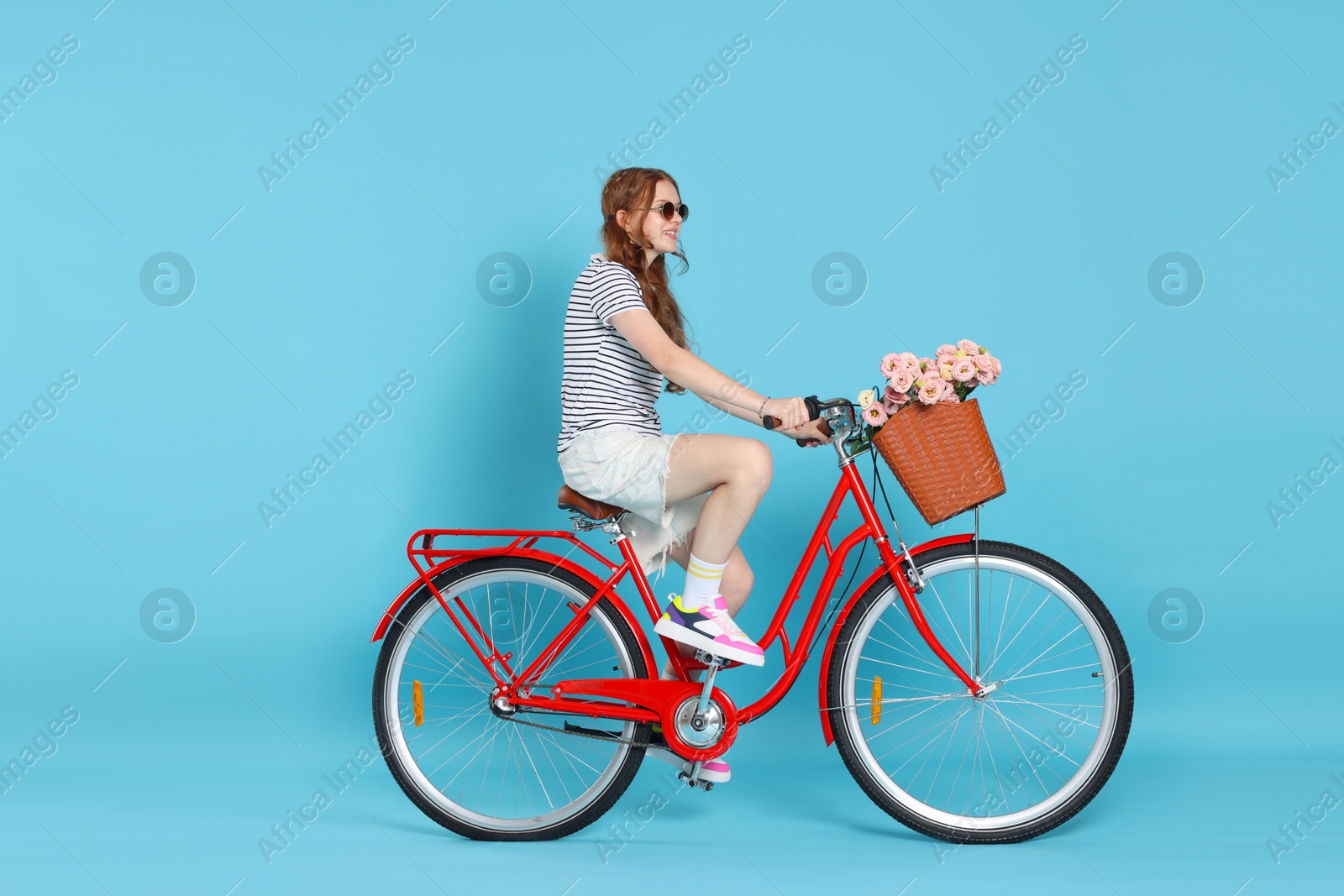 Smiling teenage girl in sunglasses riding bicycle with basket of flowers against light blue background Photo of Smiling teenage girl in sunglasses riding bicycle with basket of flowers against light blue background