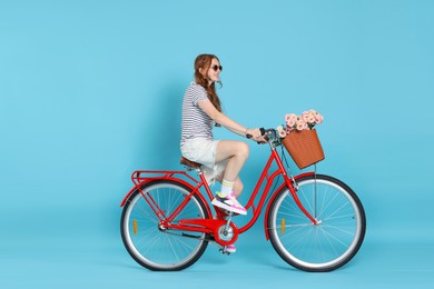Photo of Smiling teenage girl in sunglasses riding bicycle with basket of flowers against light blue background