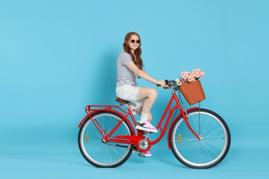 Photo of Beautiful teenage girl in sunglasses riding bicycle with basket of flowers against light blue background