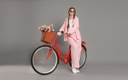 Photo of Smiling woman in sunglasses and bicycle with basket of flowers against grey background