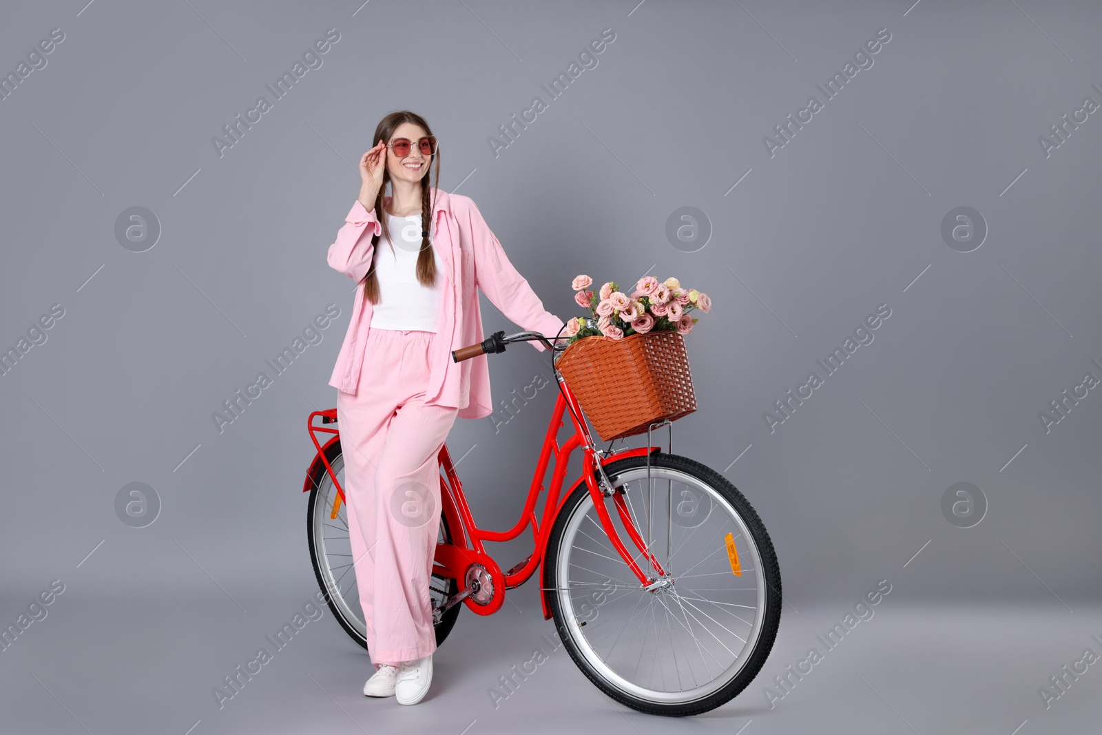 Smiling woman in sunglasses and bicycle with basket of flowers against grey background Photo of Smiling woman in sunglasses and bicycle with basket of flowers against grey background