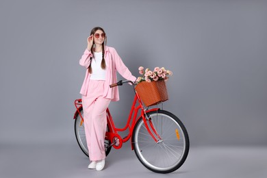 Photo of Smiling woman in sunglasses and bicycle with basket of flowers against grey background