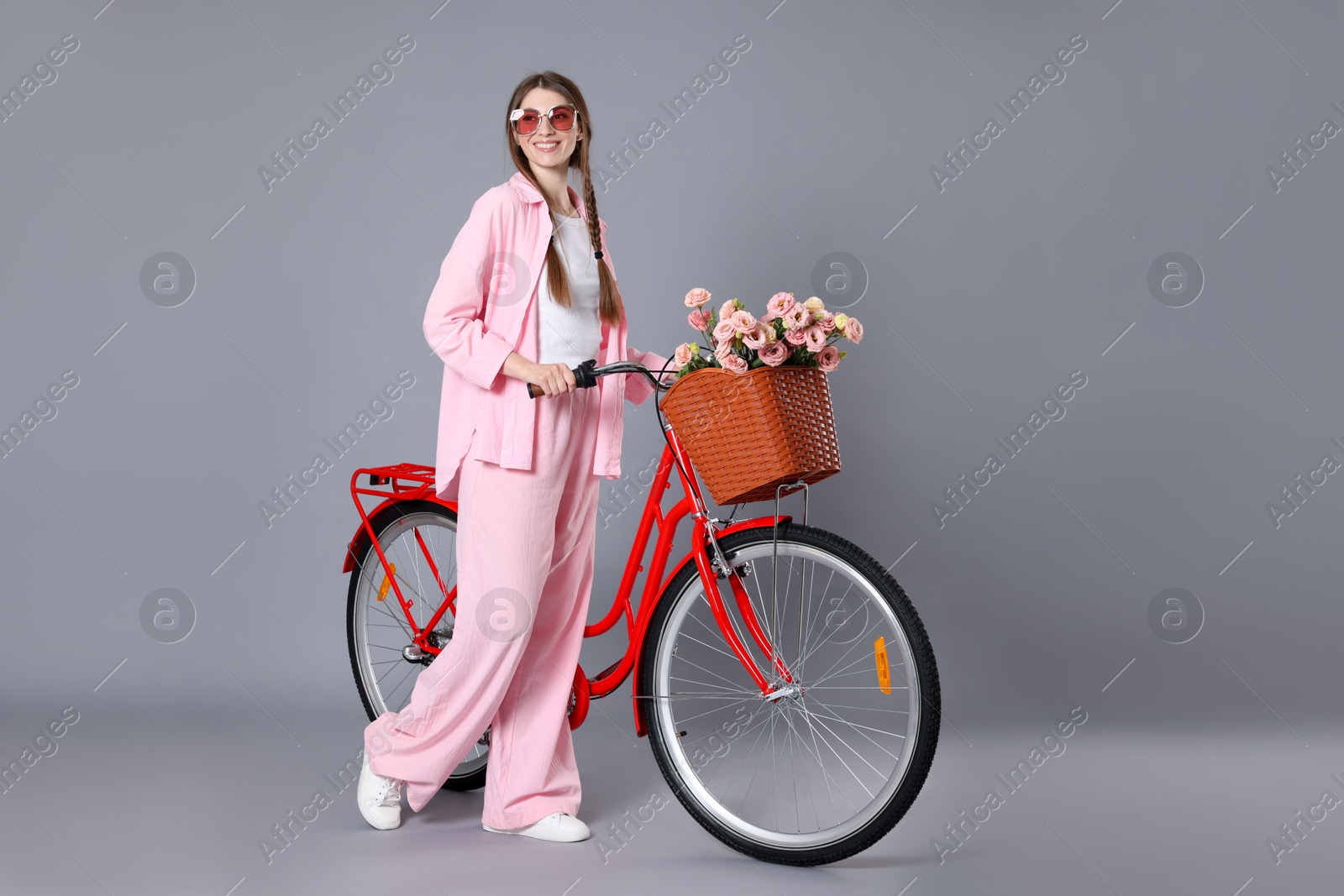 Smiling woman in sunglasses and bicycle with basket of flowers against grey background Photo of Smiling woman in sunglasses and bicycle with basket of flowers against grey background