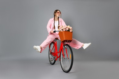 Photo of Happy woman in sunglasses riding bicycle with basket of flowers against grey background