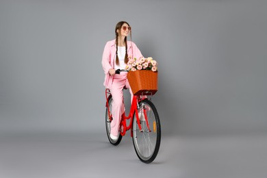 Photo of Smiling woman in sunglasses riding bicycle with basket of flowers against grey background