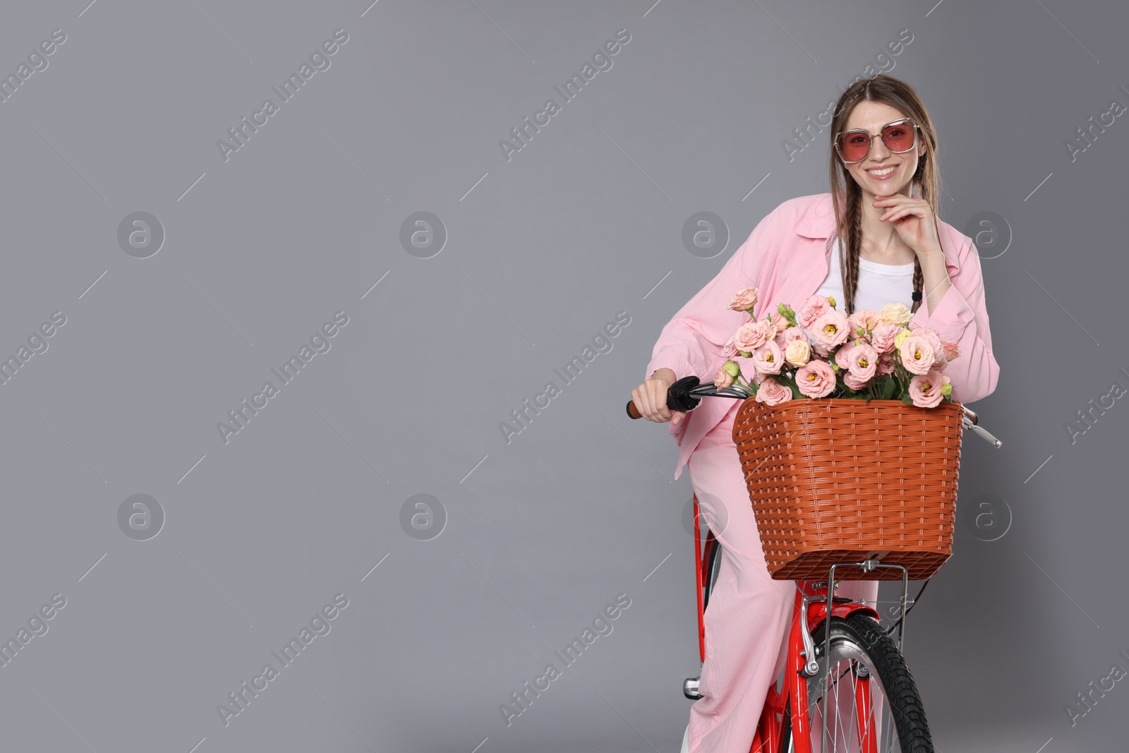 Smiling woman in sunglasses posing on bicycle with basket of flowers against grey background. Space for text Photo of Smiling woman in sunglasses posing on bicycle with basket of flowers against grey background. Space for text