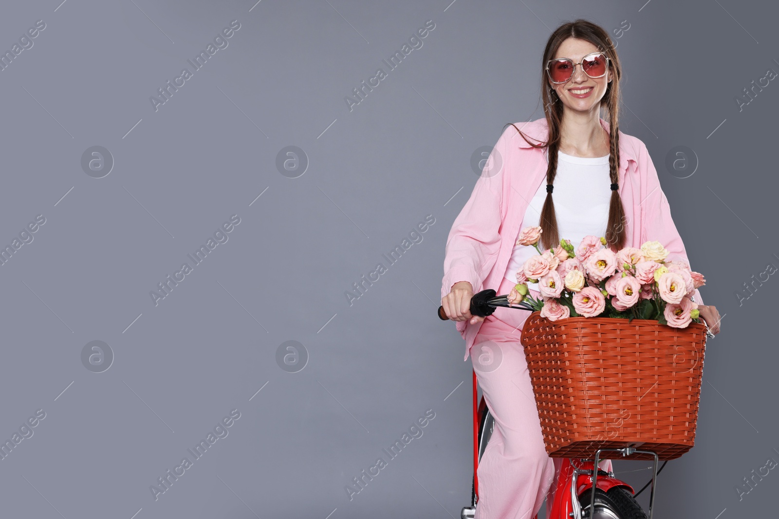 Photo of Smiling woman in sunglasses riding bicycle with basket of flowers against grey background. Space for text
