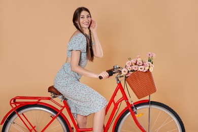 Photo of Smiling woman in dress riding bicycle with basket of flowers on beige background