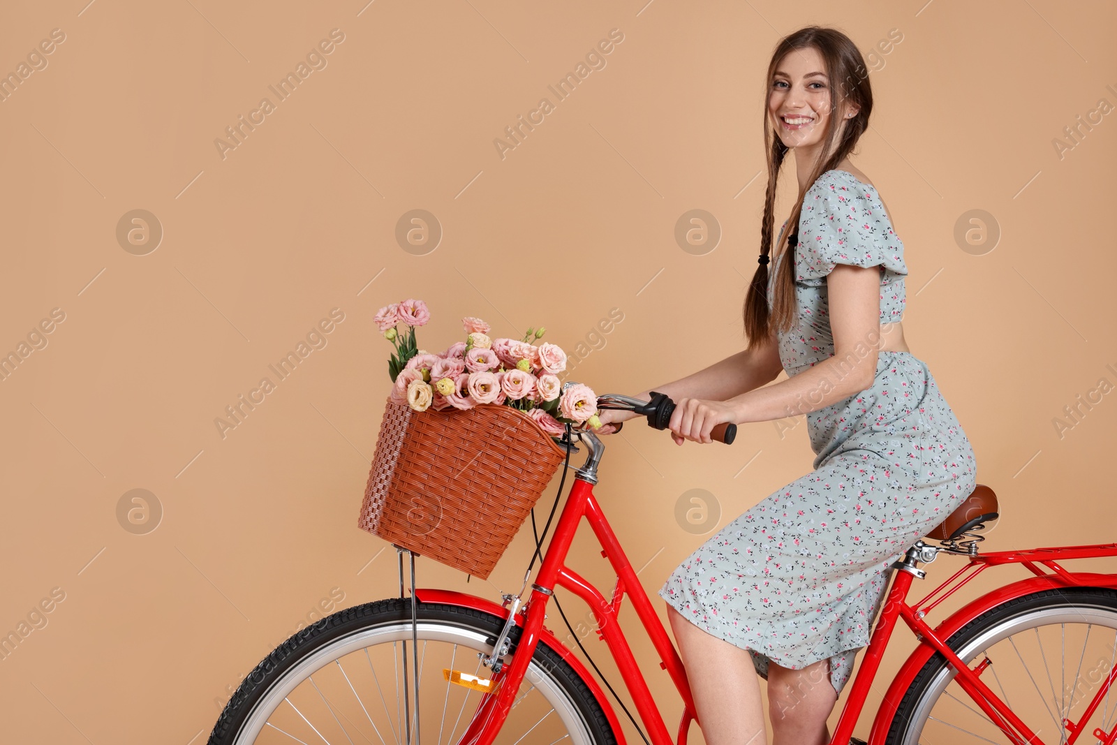 Smiling woman in dress riding bicycle with basket of flowers on beige background Photo of Smiling woman in dress riding bicycle with basket of flowers on beige background