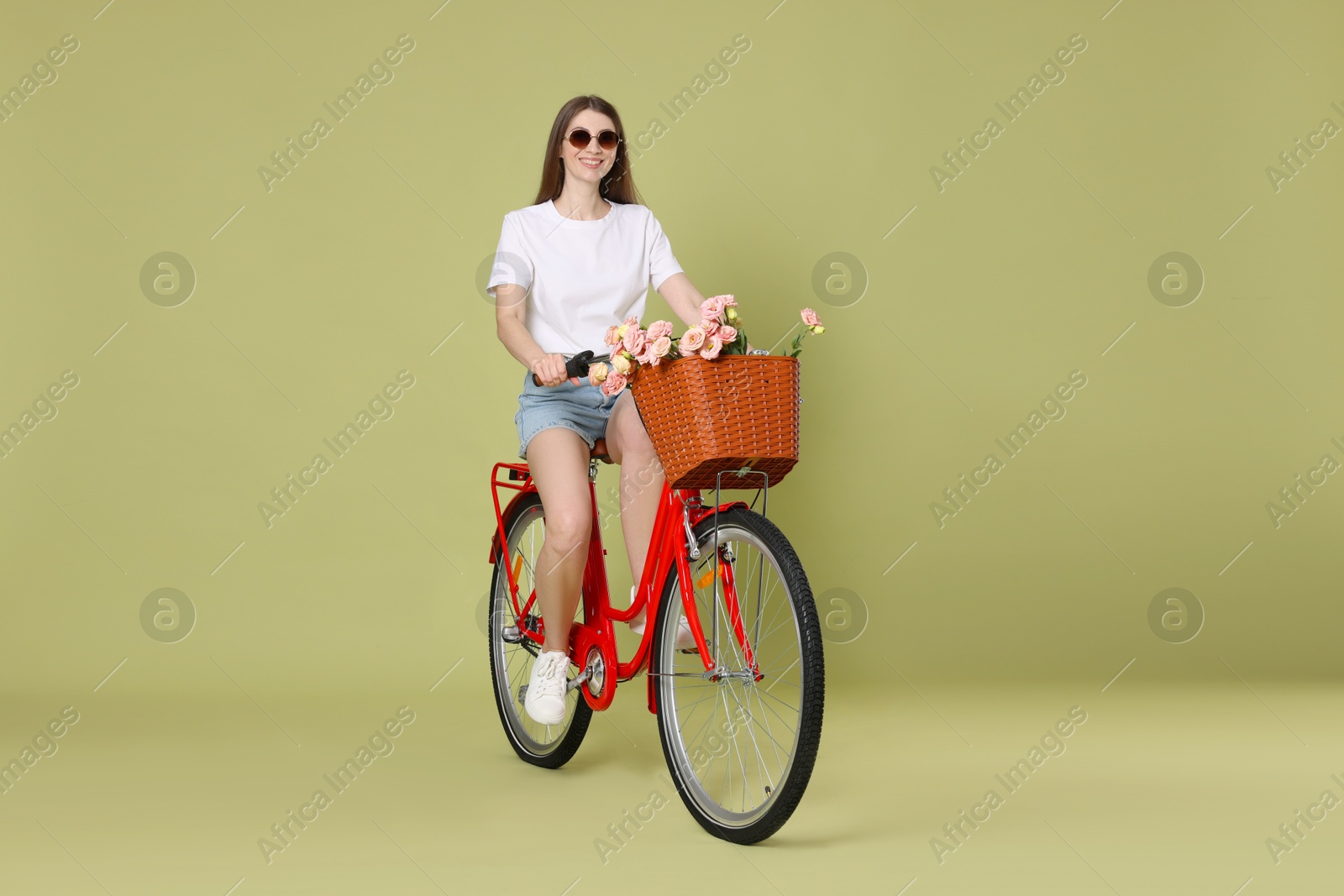 Photo of Smiling woman in sunglasses riding bicycle with basket of flowers on green background