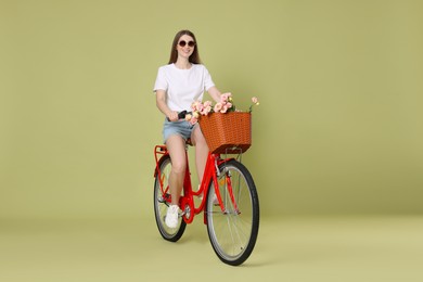 Photo of Smiling woman in sunglasses riding bicycle with basket of flowers on green background
