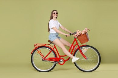 Photo of Smiling woman in sunglasses riding bicycle with basket of flowers on green background
