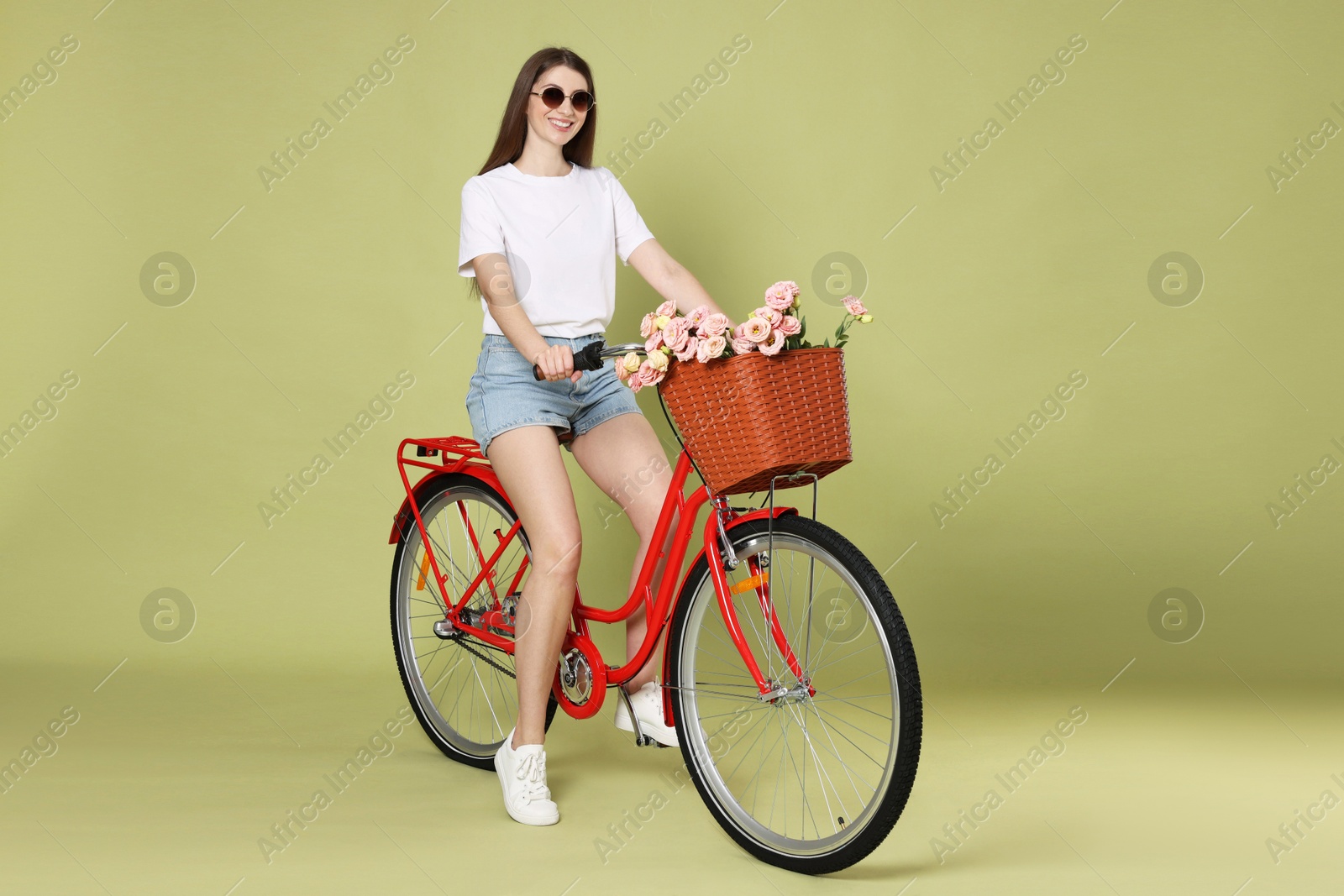 Photo of Smiling woman in sunglasses riding bicycle with basket of flowers on green background