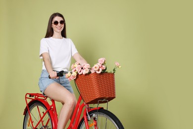 Photo of Smiling woman in sunglasses riding bicycle with basket of flowers on green background. Space for text