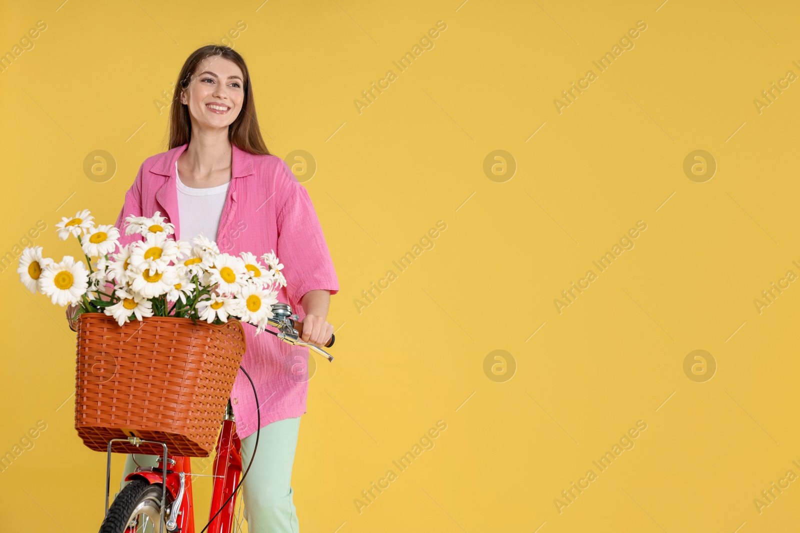 Photo of Smiling woman riding bicycle with basket of flowers against yellow background. Space for text