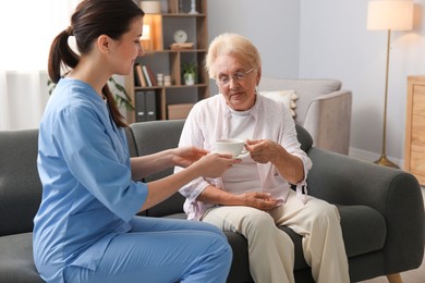 Photo of Nurse giving cup of drink to senior woman indoors. Home health care service