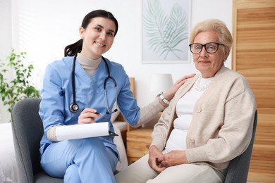 Photo of Nurse with clipboard examining senior woman indoors. Home health care service