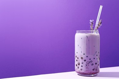 Photo of Tasty milk bubble tea in glass and lavender flowers on white table against purple background, space for text