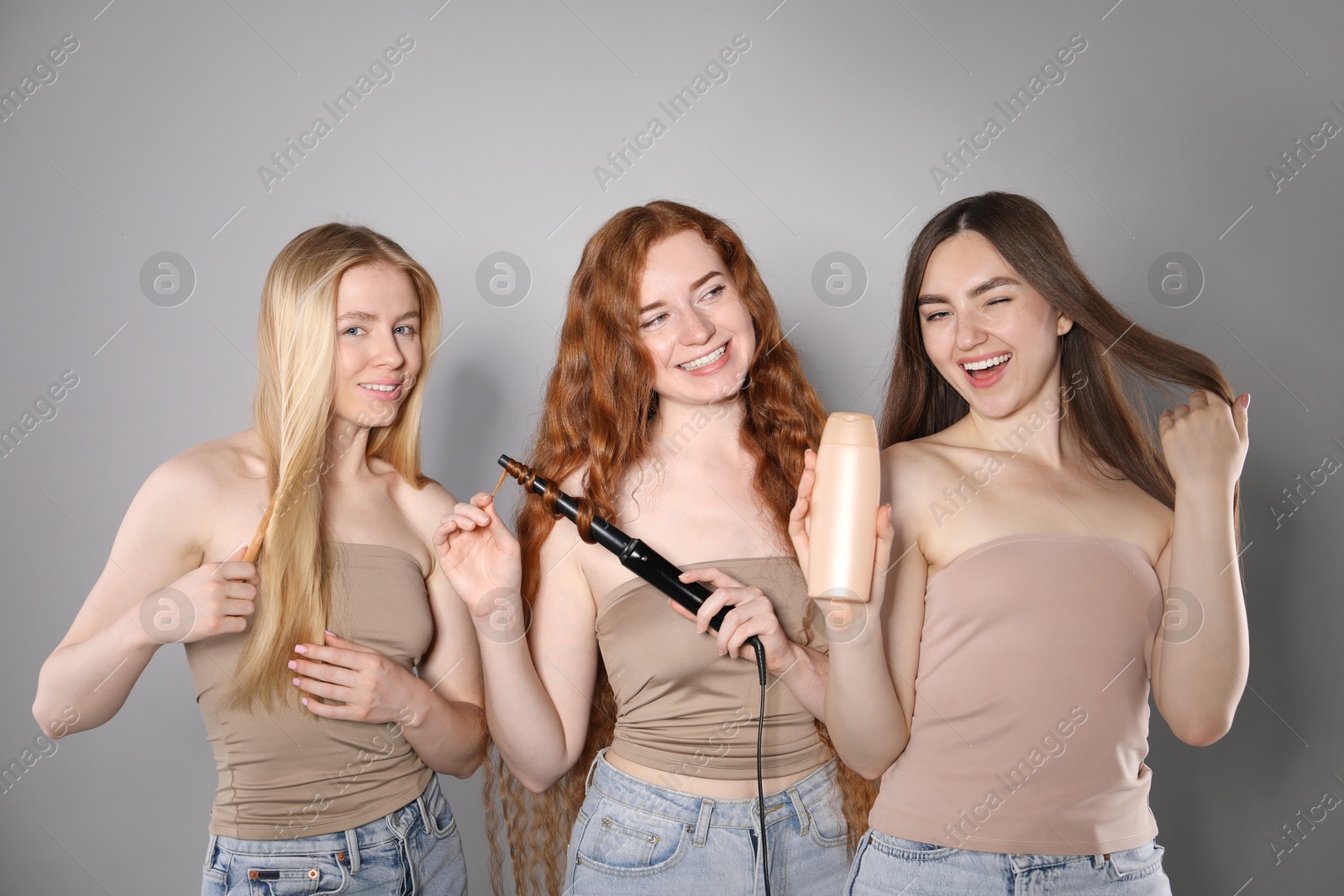 Beautiful women with different hair colors holding curling iron, shampoo and brush on grey background Photo of Beautiful women with different hair colors holding curling iron, shampoo and brush on grey background