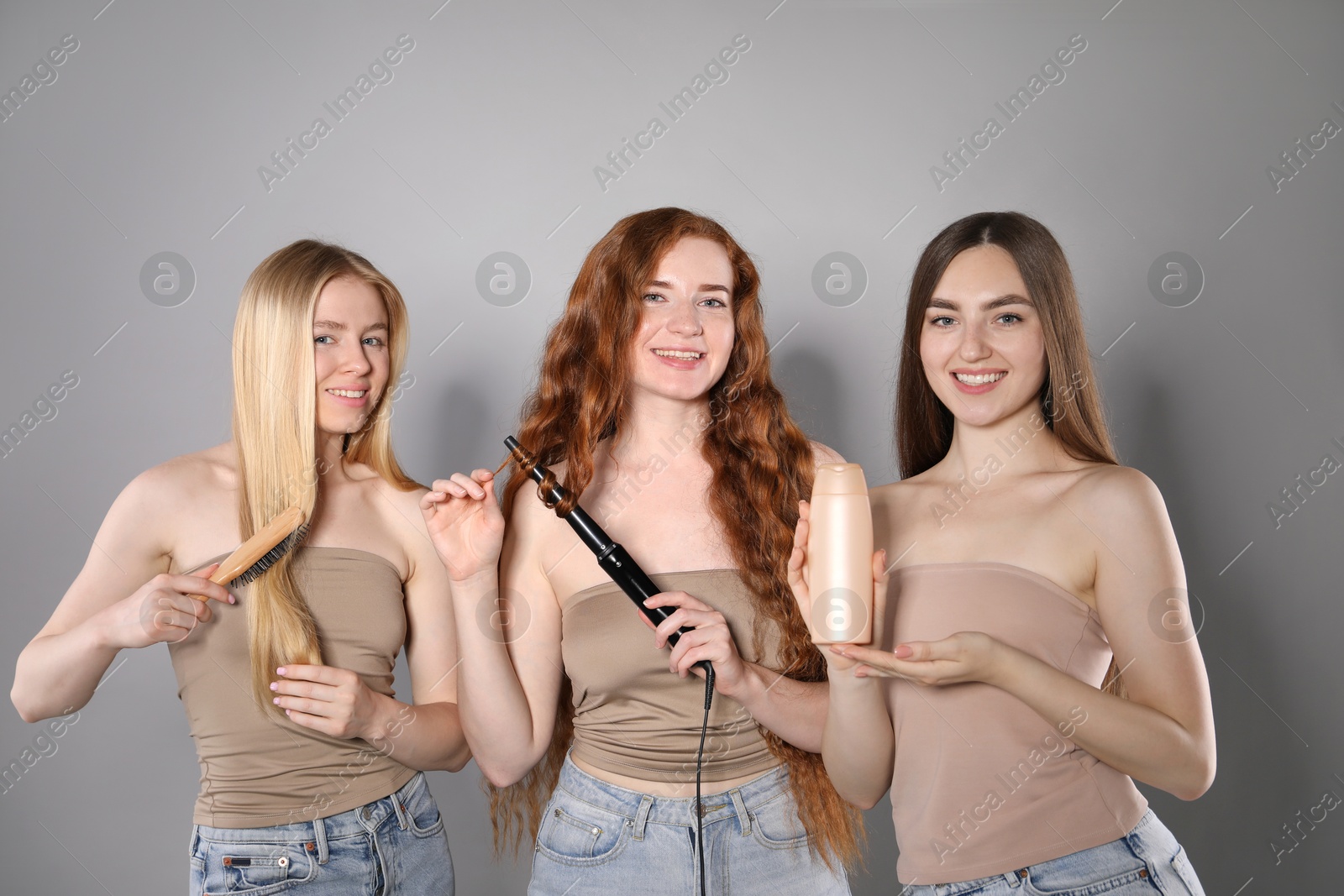 Beautiful women with different hair colors holding curling iron, shampoo and brush on grey background Photo of Beautiful women with different hair colors holding curling iron, shampoo and brush on grey background