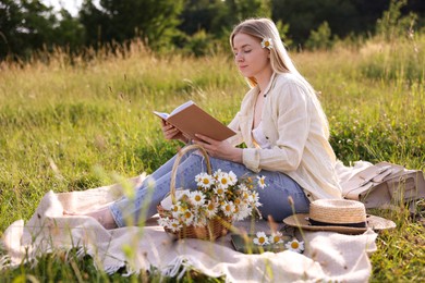Photo of Woman with bouquet of wildflowers reading book on blanket in meadow