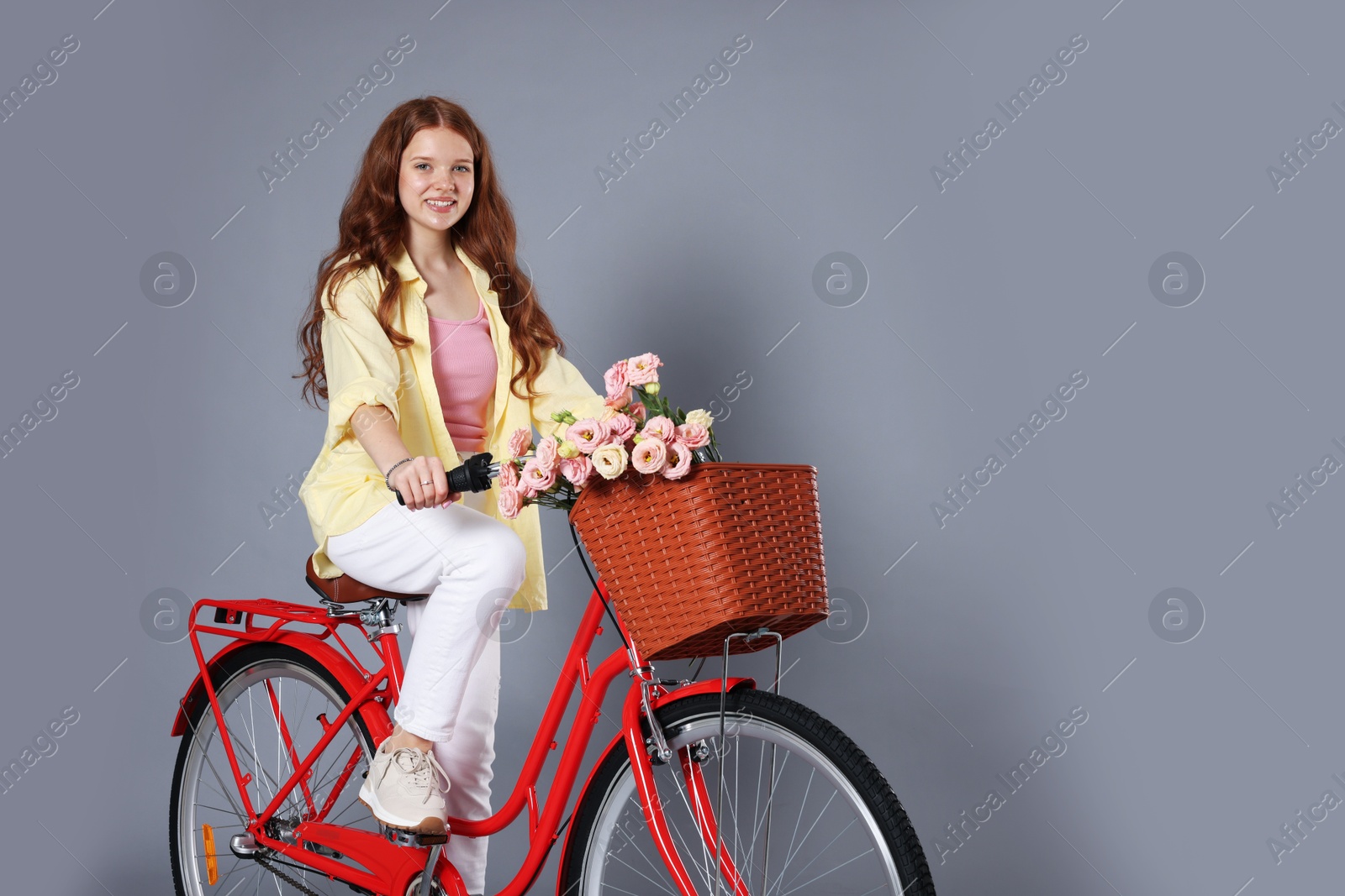 Smiling teenage girl riding bicycle with basket of flowers against grey background Photo of Smiling teenage girl riding bicycle with basket of flowers against grey background