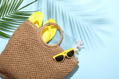 Photo of Wicker bag with beach towel, sunglasses, flip flops and palm leaf on light blue background, flat lay. Space for text